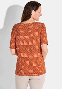 Kurze Ärmeltop in Rot mit einem geometrischen Muster aus gelben und cremefarbenen Formen. Glatte Textur und runder Ausschnitt.