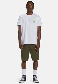 T-shirt en coton blanc avec impression graphique verte et logo, associé à un short cargo vert olive, des chaussettes blanches et des sneakers montantes noires.