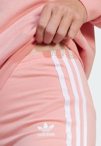 Světlé růžové sportovní oblečení s bílými pruhy po stranách a malým bílým logem. Měkká textura s priléhavým střihem.