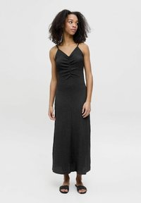 Abito maxi nero con spalline sottili, tessuto texture, dettaglio di arricciatura anteriore e orlo dritto. Abbinato a sandali neri piatti in tessuto intrecciato.