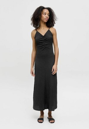 Abito maxi nero con spalline sottili, tessuto texture, dettaglio di arricciatura anteriore e orlo dritto. Abbinato a sandali neri piatti in tessuto intrecciato.
