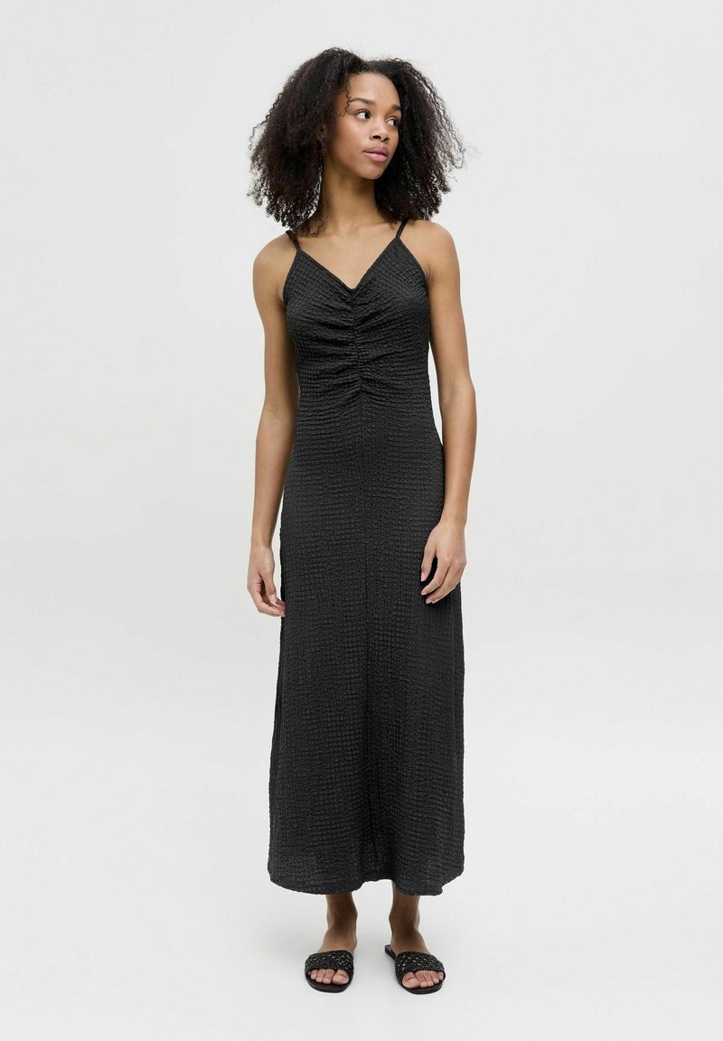 Abito maxi nero con spalline sottili, tessuto texture, dettaglio di arricciatura anteriore e orlo dritto. Abbinato a sandali neri piatti in tessuto intrecciato.
