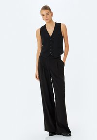 Zwarte vest met een v-hals en zes knopen, gecombineerd met hoge taille, wijde broekspijpen. Gladde stof zonder zichtbare patronen of texturen.