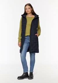 Marineblaues gepolstertes Weste mit Kapuze, Seitentaschen und Frontreißverschluss. Olivgrünes Langarmoberteil, kombiniert mit blauen Jeans und schwarzen Stiefeletten.