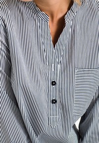 Camisa a rayas en azul marino y blanco con escote en V, dos botones y un bolsillo en el pecho. La tela parece suave con una textura ligera.