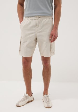 Hombre con pantalones cortos cargo beige con bolsillos y zapatillas blancas, de pie con las manos en los bolsillos frente a un fondo liso.