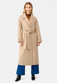 Beige wollen jas met een dubbelzijdig ontwerp, grote revers, riem in de taille en brede mouwen. Gecombineerd met blauwe wijde broek en zwarte laarzen.