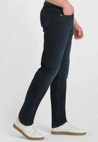 Jean en denim bleu foncé avec une coupe slim, taille mi-haute et design à cinq poches, associés à des baskets blanches et une couture latérale texturée.