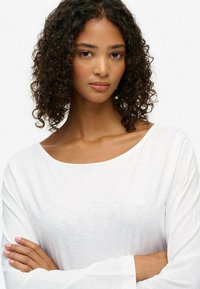 Witte lange mouwen top met een brede halslijn, met een zachte textuur en subtiele glans, gedragen door een model met krullend donker haar.