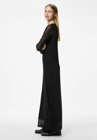 Černé šaty s dlouhým rukávem, úzkým tělem a splývavou sukní z lehkého materiálu; průhledné rukávy, kulatý výstřih a délka po kotníky.