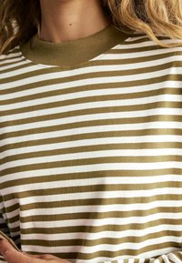 Raita pitkähihaine paita, jossa on oliivinvihreitä ja valkoisia vaakaraitoja, ribattua pyöreää kaula-aukkoa ja pehmeää puuvillamateriaalia.