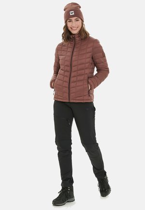 Bruine gewatteerde jas met een hoge kraag, zwarte broek en zwarte laarzen. De jas heeft een ritssluiting aan de voorkant en zijzakken.