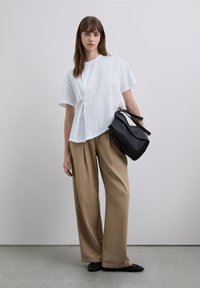 Weißes kurzärmliges T-Shirt mit einem drapierten Design, kombiniert mit locker sitzenden beigen Hosen und einer schwarzen Lederhandtasche mit strukturierten Akzenten.