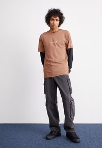 Brown cotton t-shirt with white lettering, layered over a black long-sleeve shirt. Worn with dark gray cargo pants and black sneakers.