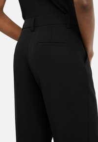 Pantaloni nero su misura con un tessuto liscio, dotati di una tasca posteriore e una silhouette snella. La vita ha passanti per una cintura.