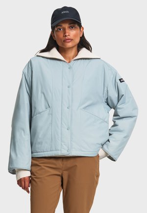 Femme portant une veste matelassée bleu clair, pantalon beige, manches de pull blanc visibles, et casquette de baseball noire, debout avec une main dans la poche.