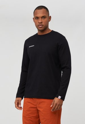 Man die een zwart shirt met lange mouwen met het "Mammut" logo draagt en een oranje broek, staand tegen een effen lichtgrijze achtergrond.
