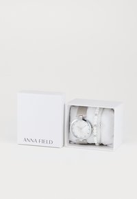 Sidabrinė tinklelio laikrodžio apyrankė su balta ciferblatu ir diskretiškais valandų žymekliais, derinama su perlų apyranke, pateikta baltame dėžutėje su "ANNA FIELD" logotipu.