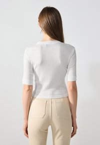 Top blanco de punto acanalado con mangas cortas, escote redondo y corte cropped, combinado con pantalones beige con bolsillos traseros y una textura suave.