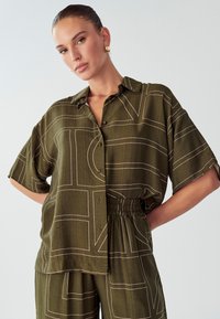 Una donna con i capelli lisciati all'indietro indossa un completo coordinato color verde oliva, con una camicia e pantaloni con linee geometriche bianche, in posa con una mano sulla vita.