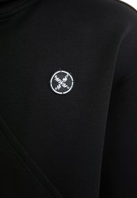 Sudadera negra con una tela suave y texturizada. Presenta un logotipo circular bordado en blanco y detalles de costura diagonal en la parte frontal.