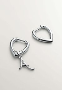 Pendientes de plata en forma de corazón con un acabado suave. Uno de los pendientes presenta un charm colgante con la letra "A".