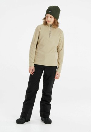 Pull en polaire beige avec demi-zip et détail de logo, associé à un pantalon imperméable noir et des bottes noires, complété par un bonnet vert.
