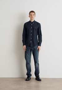 Marineblauw overhemd met witte knopen, gecombineerd met donkere denim jeans en zwarte schoenen. Het shirt heeft een klein logo op de borst.