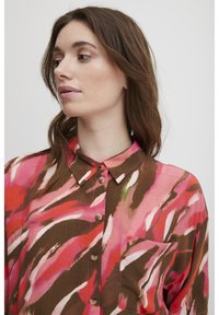 Camicia multicolore con un motivo astratto rosa e marrone, colletto, chiusura frontale con bottoni e taschino sul petto. Realizzata in tessuto leggero.