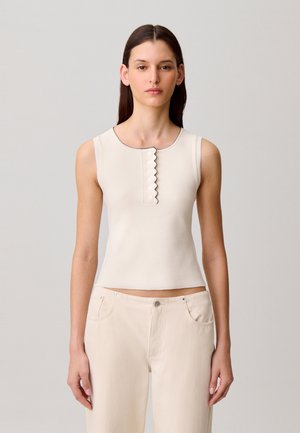 Haut sans manches crème avec un col arrondi, doté d'un détail festonné et d'une patte de boutonnage à cinq boutons. Tissu lisse et extensible avec une coupe ajustée.