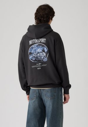 Personne portant un sweat à capuche noir avec un graphisme de sport automobile et une illustration de moto dans le dos, associé à un jean bleu ample.