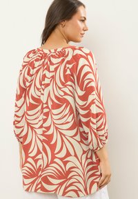 Frau in einer weiten, langärmeligen Bluse mit rot-beigem abstraktem Blattmuster, von hinten vor einem schlichten hellen Hintergrund betrachtet.