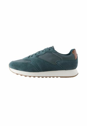 Mørkegrøn fløjl og stof sneakers med brun hælflap, hvid sål og struktureret tan ydersål vist i sideprofil.