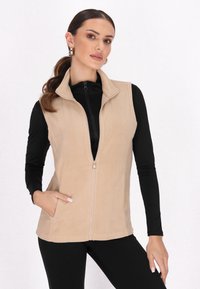 Beige Fleecepullover mit Reißverschluss, Stehkragen und Seitentaschen. Über einem schwarzen Langarmoberteil getragen. Glatte Textur, lässiges Design.