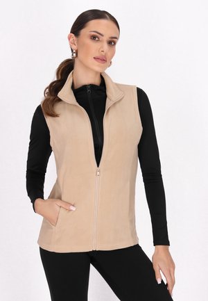 Gilet in pile beige con zip frontale, colletto alto e tasche laterali. Indossato sopra una maglietta nera a maniche lunghe. Texture liscia, design casual.