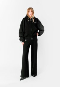 Sudadera negra oversize con cremallera y detalle de anillo, combinada con pantalones de campana negros. Presenta detalles de logo y una silueta relajada.