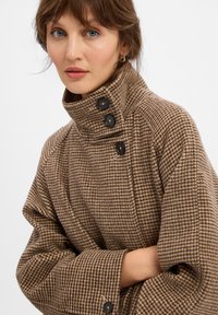 Frau mit blauen Augen, die einen braun-beige Karomantel mit hohem Kragen und schwarzen Knöpfen trägt, mit verschränkten Armen.