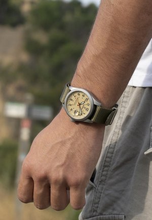 Zilveren horloge met crème wijzerplaat, zwarte uurmarkeringen en rode accenten. Oliijfkleurige stoffen band. Zichtbare hand rustend in een vuist.