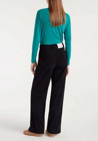 Una blusa de manga larga en color turquesa y pantalones de pierna ancha negros con una etiqueta de cinturilla visible, combinados con calzado beige. Textura de tela suave.