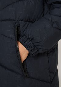 Detail na tmavě modré prošívané bundě ukazující zipovou kapsu, elastické manžety a strukturovaný vzor na hladkém materiálu.