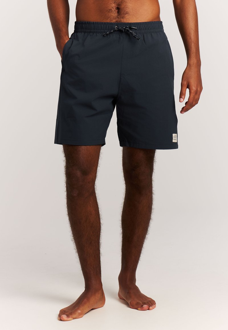 Short de bain sombre avec taille élastique et cordon porté par un homme pieds nus debout devant un fond neutre.