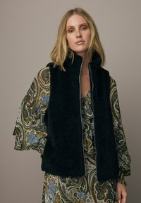 Černá fleecová vesta na zip s vysokým límcem; nošena přes vzorované, volné šaty s modrými, zelenými a zlatými květinovými vzory.