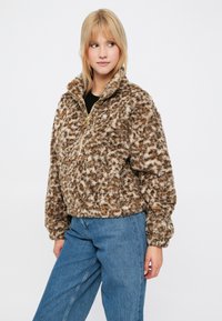 Brązowy pulower z sztucznego futra z wysokim kołnierzem, ćwierć-zamkiem z przodu oraz teksturowanym, cętkowanym wzorem. Połączony z niebieskimi dżinsami.