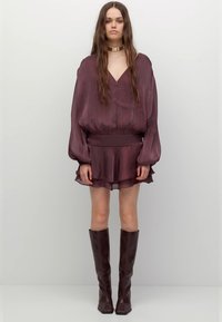 Blusa color prugna scuro con maniche lunghe, scollo a V e vita arricciata. Abbinate a una minigonna a strati e stivali marroni al ginocchio con punta quadrata.