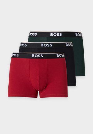 Homme musclé debout pieds nus portant un boxer rouge BOSS, avec un bras croisé sur la poitrine sur un fond blanc uni.