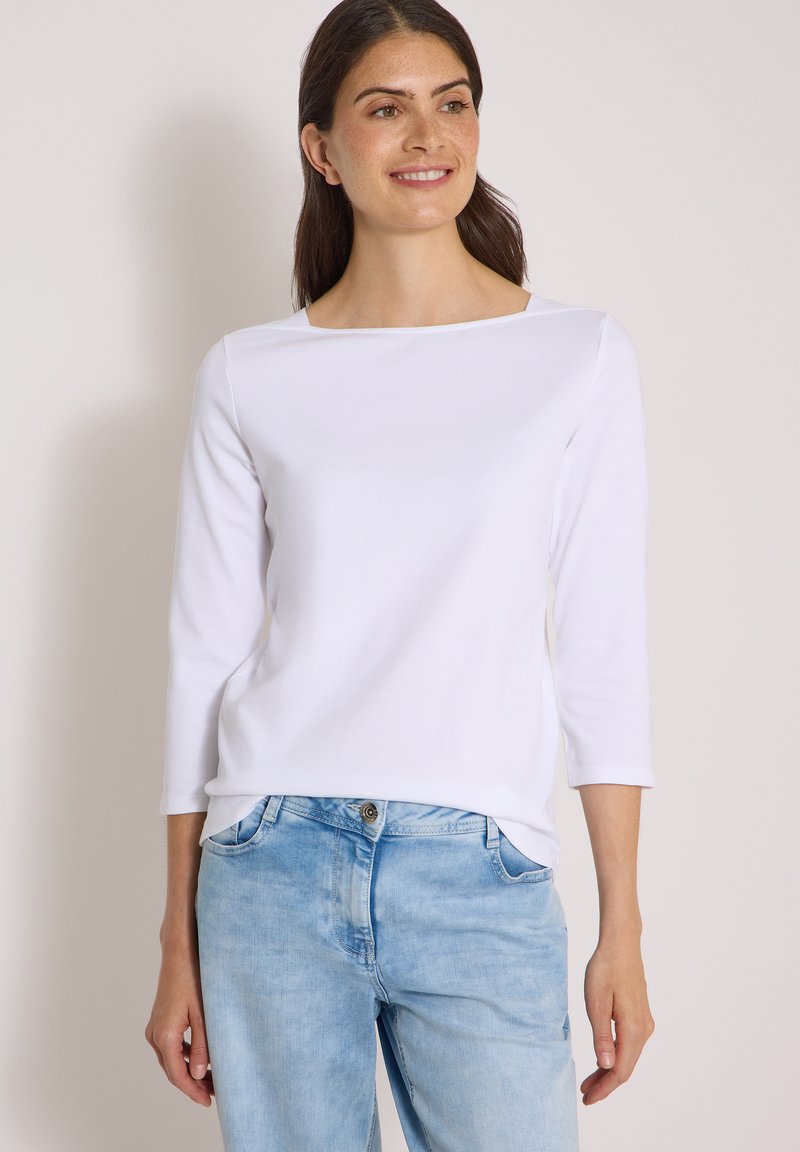 Femme portant un haut blanc à manches longues et un jean bleu clair, debout devant un fond clair uni, souriante et regardant légèrement vers la droite.