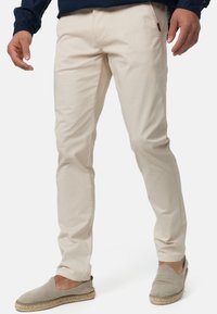 Chino beige en coton avec une coupe slim. Dispose de poches avant et d'une étiquette logo discrète sur le côté. Porté avec des chaussures slip-on marron.