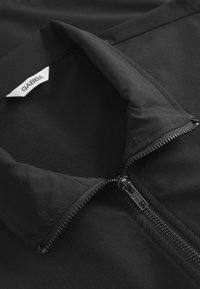 Melns džemperis ar rāvējslēdzēja apkakli un uzlīmi ar uzrakstu "GABBA." Raksturo gluda tekstūra un izturīgas rāvējslēdzēja detaļas, galvenokārt tumšās krāsās.