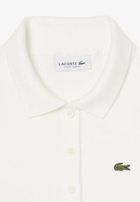 Biała koszulka polo Lacoste o fakturze pikowanej, w wąskim kroju, z prążkowanym kołnierzykiem oraz zielonym haftowanym logo krokodyla w pobliżu górnego lewego guzika.