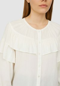 Camicia bianca con design a volant al colletto e alle spalle, caratterizzata da una chiusura con bottoni sul davanti e realizzata in un tessuto liscio e leggero.